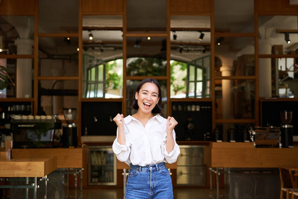 Χαρούμενη ενθουσιώδης Ασιάτισσα, ιδιοκτήτρια επιχείρησης θριαμβεύει, ανοίγει το δικό της εστιατόριο, στέκεται μπροστά σε άδειο καφέ.. - Φωτογραφία, εικόνα