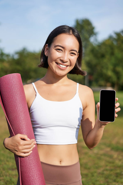 Pionowy strzał zdrowej i zdrowej młodej azjatyckiej kobiety, pokazuje ekran smartfona ze statystykami treningu, zaleca aplikację telefoniczną. - Zdjęcie, obraz