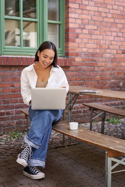 Вертикальный снимок азиатки, работающей в кафе на открытом воздухе, сидящей с ноутбуком на скамейке и пьющей кофе. Копирование пространства - Фото, изображение