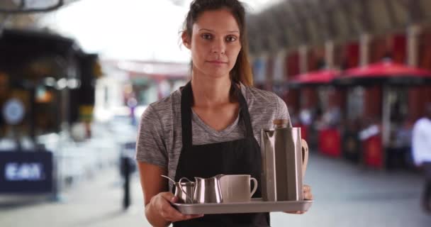 Unhappy waitress holding tray with coffee and tea outside. Portrait of serious attractive woman wearing apron and carrying tray to serve beverages. 4k - Footage, Video