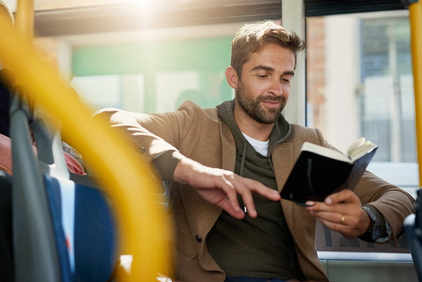 Του αρέσει να διαβάζει όσο είναι στο λεωφορείο. Ένας όμορφος νεαρός διαβάζει ένα βιβλίο κατά τη διάρκεια της πρωινής του μετακίνησης με λεωφορείο. - Φωτογραφία, εικόνα