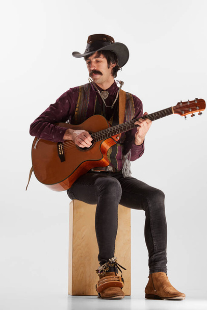 Portrait of man with moustaches in country style clothes playing guitar isolated over white background. Concept of music, creativity, inspiration, hobby, lifestyle, live performance - Foto, Bild