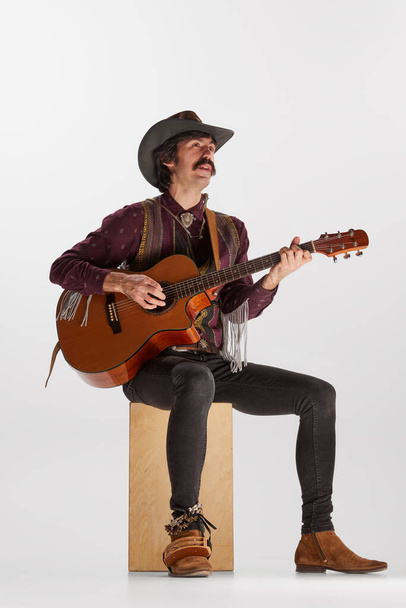 Portrait of man with moustaches in country style clothes playing guitar, singing isolated over white background. Performer. Concept of music, creativity, inspiration, hobby, lifestyle - Photo, Image