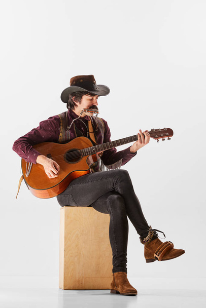 Portrait of man with moustaches in country cowboy style clothes playing guitar and harmonica, performing isolated over white background. Concept of music, creativity, inspiration, hobby, lifestyle - Photo, Image