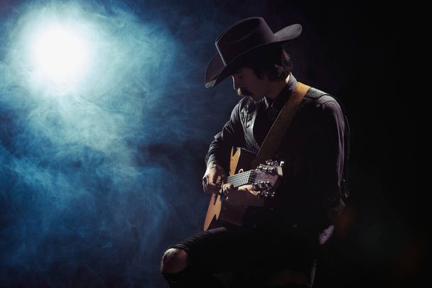 Portrait of man with moustaches in country style clothes playing guitar isolated over dark blue background with smoke. Night performer. Concept of music, creativity, inspiration, hobby, lifestyle - Zdjęcie, obraz