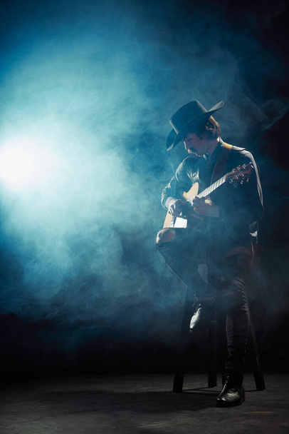 Portret van de mens met snorren in country style kleding die gitaar speelt geïsoleerd over donkerblauwe achtergrond met rook. Jazz artiest. Concept muziek, creativiteit, inspiratie, hobby, levensstijl - Foto, afbeelding