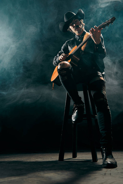 Portrait of man with moustaches in country style clothes playing guitar isolated on dark blue background with smoke. Expression of feelings. Concept of music, creativity, inspiration, hobby, lifestyle - Foto, afbeelding