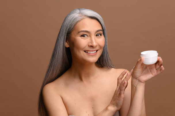 Beauty portrait of charming mature gray-haired Asian woman applying moisturising cream on hand and look at camera, doing an allergy test, isolated over brown background. Skin care concept - Fotografie, Obrázek