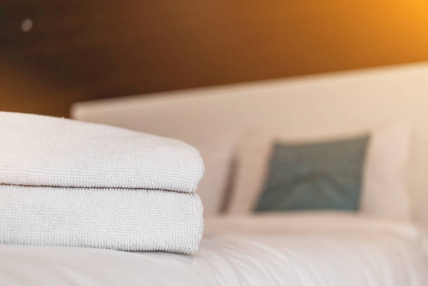 Οι καθαρές λευκές πετσέτες είναι διπλωμένες δίπλα στο κρεβάτι για να είναι έτοιμες για χρήση από τους πελάτες όταν έρθει η ώρα να κάνουν ντους, επειδή οι πετσέτες είναι απαραίτητες για να μείνουν οι επισκέπτες. Θολή φόντο με Copy Space για το σχεδιασμό. - Φωτογραφία, εικόνα