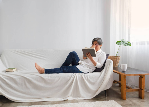 Libro de lectura de anciana inteligente con sensación de relax en el sofá en la sala de estar, estilo de vida de mediana edad - Foto, imagen