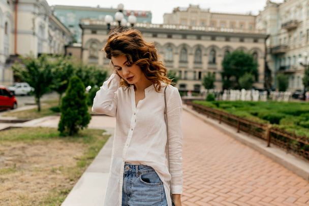 Evropská stylová mladá běloška tráví svůj volný čas o víkendu ve starém evropském městě. Nosí bílou košili a džínové kraťasy. Koncept životního stylu - Fotografie, Obrázek