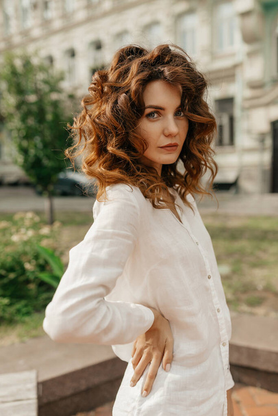 Slim attractive woman with curly red hair wearing white shirt posing outdoor on city blurred background. Nice caucasian young lady enjoys walk in nature on summer day.  - Fotografie, Obrázek