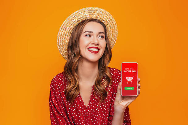 девушка держит мобильный телефон и делать онлайн-покупки изолированы на оранжевом фоне в студии - Фото, изображение