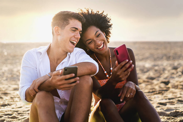 Gün batımında arka planda akıllı telefon kullanan çok ırklı bir çift gülümsüyor ve birlikte eğleniyorlar. - Fotoğraf, Görsel