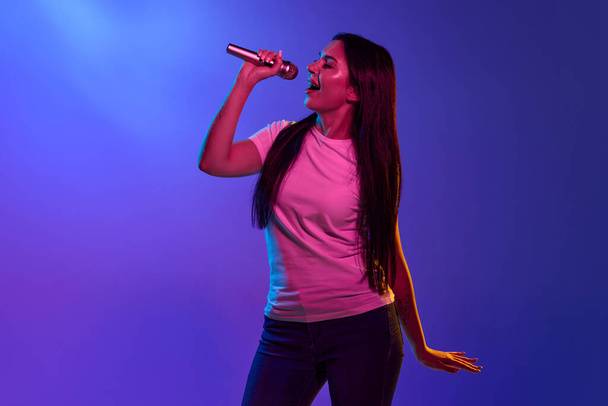 Caucasian female portrait isolated on blue studio background in pink neon light. Beautiful female model singing a song with microphone. Concept of human emotions, facial expression, ad, music, art. - Foto, imagen