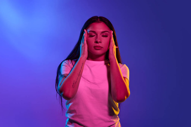 Woman having headache. Isolated on blue neon background. Girl standing with pain isolated on trendy studio background. Female half-length portrait. Human emotions, facial expression concept. - Photo, image