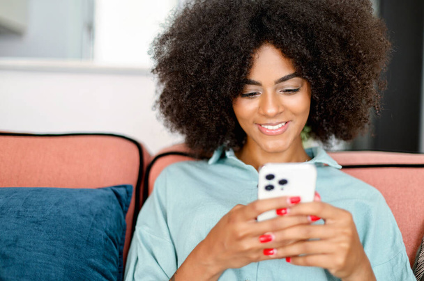 Serene young cheerful black girl positive smile sitting in relaxed pose on the couch at home and chatting online on the cellphone, replying sms, web browsing - Фото, зображення