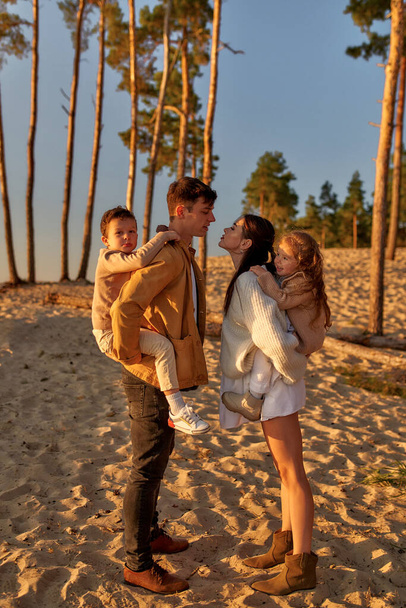 Молода сім'я з двома дітьми на спині цілується на піщаному пляжі на тлі сосен на заході сонця у вихідні, розслабляючись і розважаючись
. - Фото, зображення