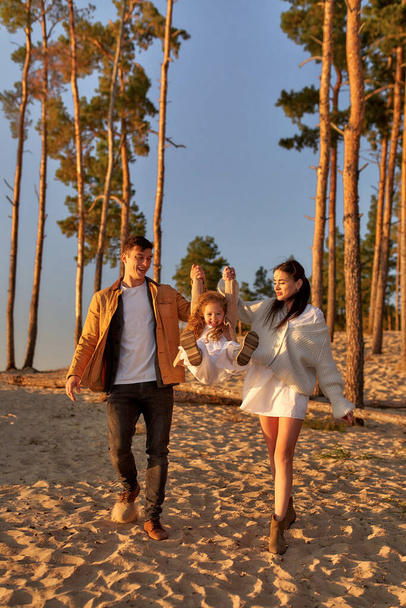 Młoda rodzina z uroczą córką trzymającą się za ręce na piaszczystej plaży na tle sosen o zachodzie słońca podczas weekendowego relaksu i zabawy. - Zdjęcie, obraz