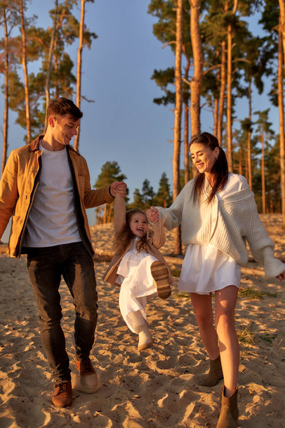 A young family with a cute daughter holding hands on a sandy beach against the backdrop of pine trees at sunset over the weekend relaxing and having fun. - Фото, зображення