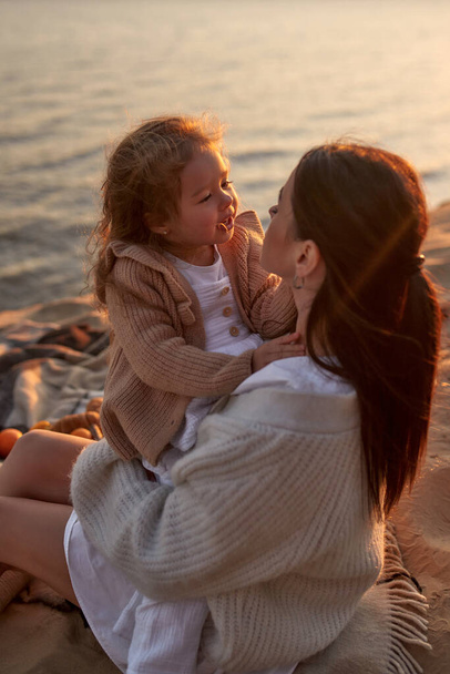 Malá roztomilá dívka v šatech a pletený svetr s její matkou sedí na dece na písečném břehu u moře při západu slunce baví a relaxaci o víkendu. - Fotografie, Obrázek