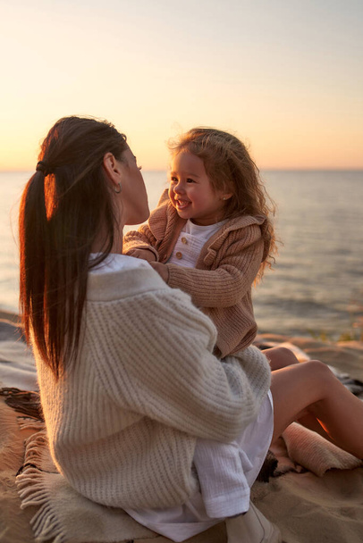 Little cute girl in a dress and a knitted sweater with her mother sitting on a blanket on a sandy shore by the sea at sunset having fun and relaxing on the weekend. - Photo, image