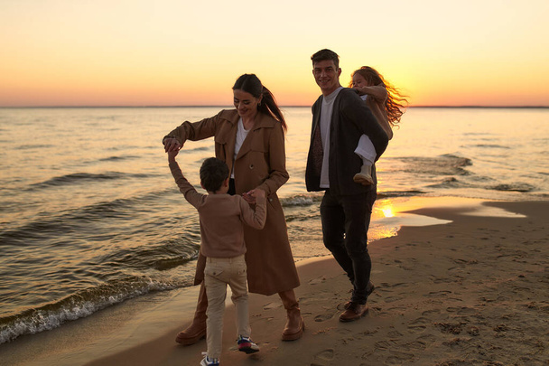 Νεαρή οικογένεια με δύο παιδιά που περπατούν στην αμμώδη παραλία κοντά στη θάλασσα το ηλιοβασίλεμα διασκεδάζοντας και παίζοντας. - Φωτογραφία, εικόνα