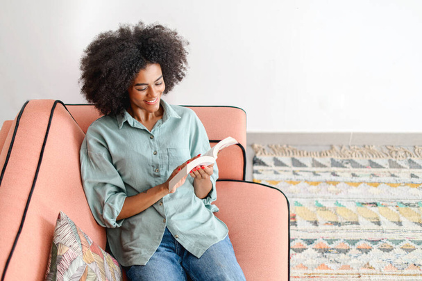 Serene young black woman enjoying reading book lying down on the comfy sofa at cozy living room, positive peaceful girl spends calm weekend at home, hobby and lifestyle concept - Foto, afbeelding
