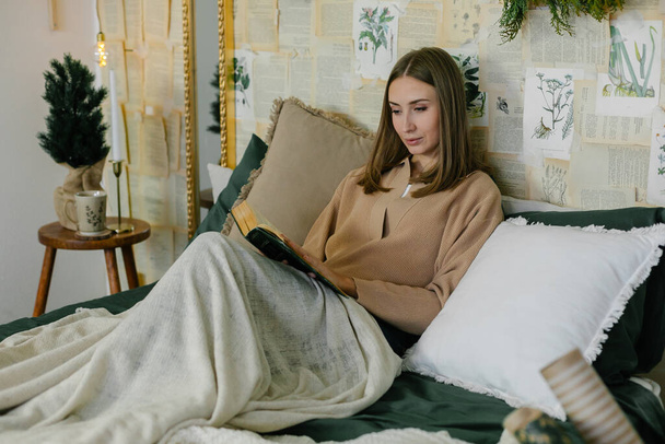 Νεαρή χαλαρή γυναίκα ανάγνωση βιβλίο, ενώ κάθεται στο κρεβάτι, χαλαρώνοντας στο σπίτι κατά τη διάρκεια των χειμερινών διακοπών. Έννοια του ζεστού χειμώνα στο σπίτι, hygge. Οικιακός τρόπος ζωής έννοια - Φωτογραφία, εικόνα