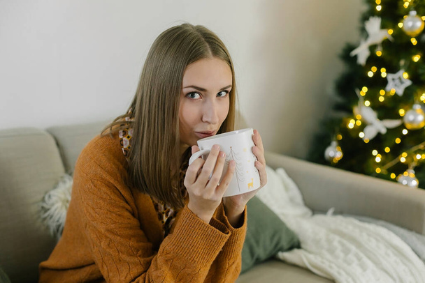 Portrait of woman relaxing at home sitting on couch near Christmas tree, drinking tea or coffee. Concept of home holiday lifestyle - Photo, Image