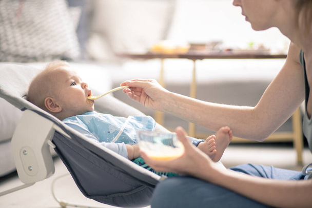 Mother spoon feeding her baby boy child in baby chair with fruit puree. Baby solid food introduction concept - Photo, Image