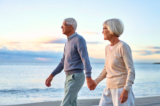 Starsza, inteligentna para zakochanych spędza romantycznie czas na plaży nad morzem, spacerując trzymając się za ręce i delikatnie przytulając. - Zdjęcie, obraz