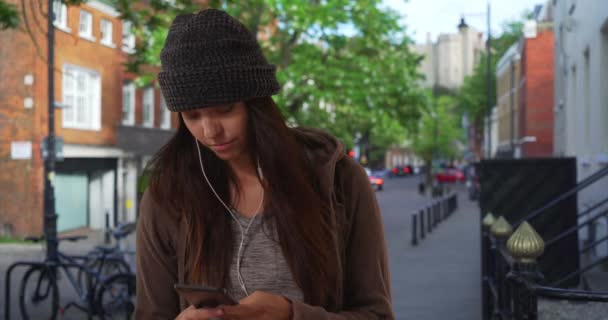 Woman on urban neighborhood street listening to music streaming app and texting with cellphone. Millennial girl in the city using mobile phone and earbuds. 4k - Footage, Video