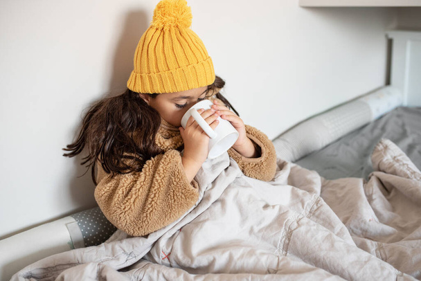 Beautiful little girl warms in blanket in cold winter time. Child in warm clothes sitting in a bed drinking hot tea at home. Cold home. No heating. - Foto, Imagem