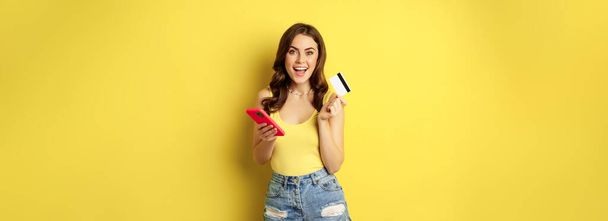 Online shopping. Stylish brunette woman holding smartphone and credit card, paying in app, using mobile phone application, buying smth, standing over yellow background. - Foto, imagen