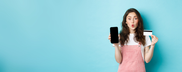 Portrét mladé krásné ženy ukazující plastové kreditní karty a prázdné obrazovky smartphone, vypadat pobaveně a zájem, ukazuje zajímavé aplikace, stojící nad modrým pozadím. - Fotografie, Obrázek