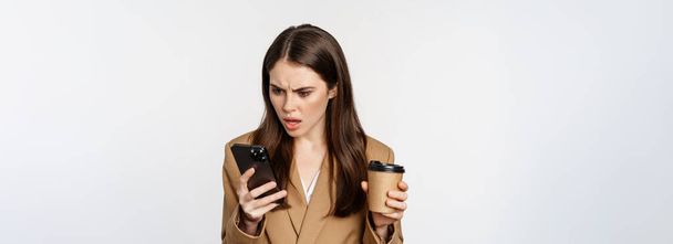 sfrustrowana kobieta biznesu wyglądająca na zszokowaną telefonem komórkowym, trzymająca kawę na wynos, stojąca nad białym tłem. - Zdjęcie, obraz