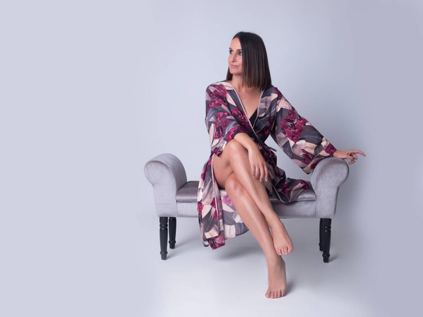 Full body barefoot female in floral silk robe looking away while sitting on soft recamier sofa during skin care routine against gray background - Zdjęcie, obraz