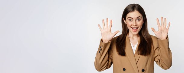 Enthusiastic businesswoman in suit, raising hands palms up, number ten dozen gesture and smiling, standing over white background. - Photo, Image