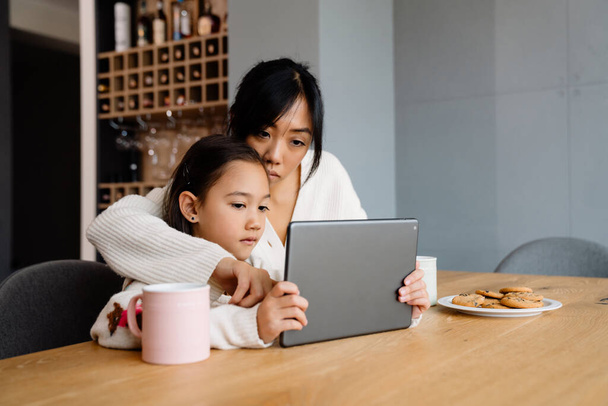Asian woman and her daughter using tablet computer while sitting at table in home - Fotografie, Obrázek