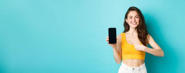 Mulher atraente feliz mostrando propaganda na tela do smartphone, apontando para a exibição vazia do telefone e sorrindo, de pé sobre o fundo azul. - Foto, Imagem