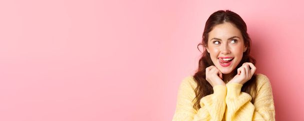 Young devious girl licking her lips and teeth, looking aside with thoughtful face, having interesting idea, thinking, standing against pink background. - Photo, Image