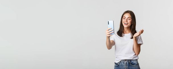 Freundlich attraktives Mädchen, das Freundin anruft, lächelt und Gespräche führt, Smartphone in der Hand, weißer Hintergrund. - Foto, Bild