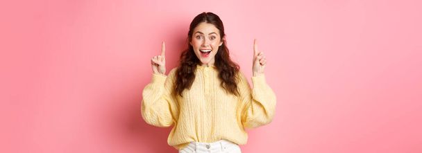 広告を表示する陽気な若い女性,プロモーションテキストで指を指して、興奮笑みを浮かべて,ピンクの背景に立って. - 写真・画像