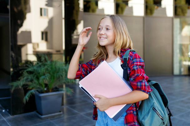Schattig 13 jaar meisje student tiener met een rugzak houdt notitieblokken en notitieboekjes op straat, glazen gebouw op de achtergrond - Foto, afbeelding