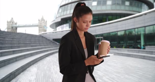 Millennial empresaria bebiendo té y mensajes de texto en el teléfono móvil. Mensajería de texto de mujer profesional joven y sosteniendo papel para ir taza de café en el parque de oficinas de negocios. 4k - Imágenes, Vídeo