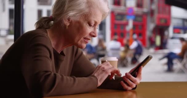 Happy senior vrouw winkelen online met creditcard en telefoon in de coffeeshop. Lachende oudere vrouw boeken vakantie op mobiele telefoon. 4k - Video