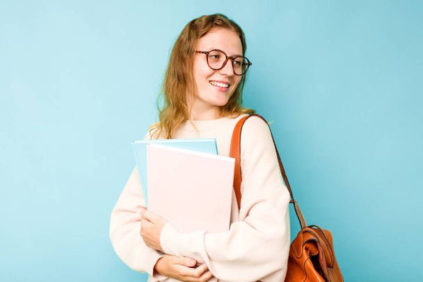 Молода студентка біла жінка ізольована на синьому фоні виглядає вбік усміхненою, веселою і приємною
. - Фото, зображення