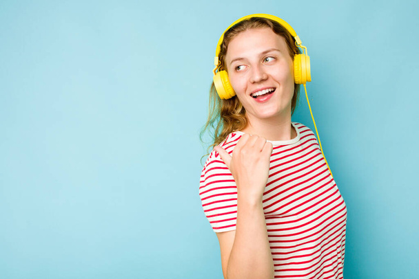 Young caucasian woman wearing headphones isolated on blue background points with thumb finger away, laughing and carefree. - Fotografie, Obrázek