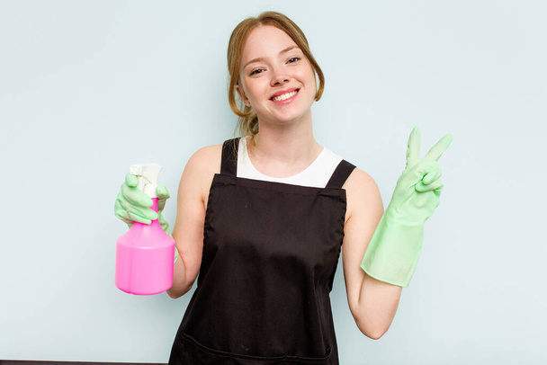 Young cleaner woman isolated on blue background joyful and carefree showing a peace symbol with fingers. - Photo, Image
